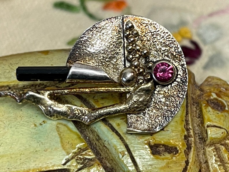 May include: A silver-toned brooch with a textured, semi-circular design. It features a black rectangular stone, a cluster of small silver spheres, and a round, pink gemstone. The brooch has a branch-like element extending from the base.