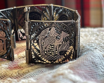 May include: A silver bracelet with a filigree design featuring a camel and rider. The bracelet is made of multiple linked sections and has a spring clasp.