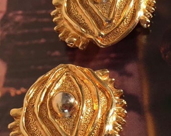 May include: A pair of gold-tone earrings with a textured, abstract design. Each earring features a raised, circular center piece.