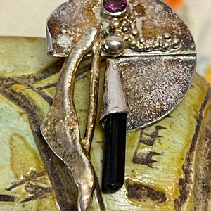 May include: A silver-toned brooch with a circular design, featuring a small, round, purple gemstone and a black, elongated stone. The brooch has an abstract, organic shape with textured and smooth surfaces, and gold-colored accents.