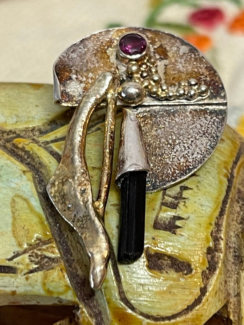 May include: A silver-toned brooch with a circular design, featuring a small, round, purple gemstone and a black, elongated stone. The brooch has an abstract, organic shape with textured and smooth surfaces, and gold-colored accents.
