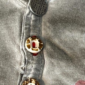 May include: Close-up of a gray ribbed knit shirt with three buttons. The top button is dark gray with embossed text. The other two buttons are white with brown and tan patterns. The shirt has a V-neck design.