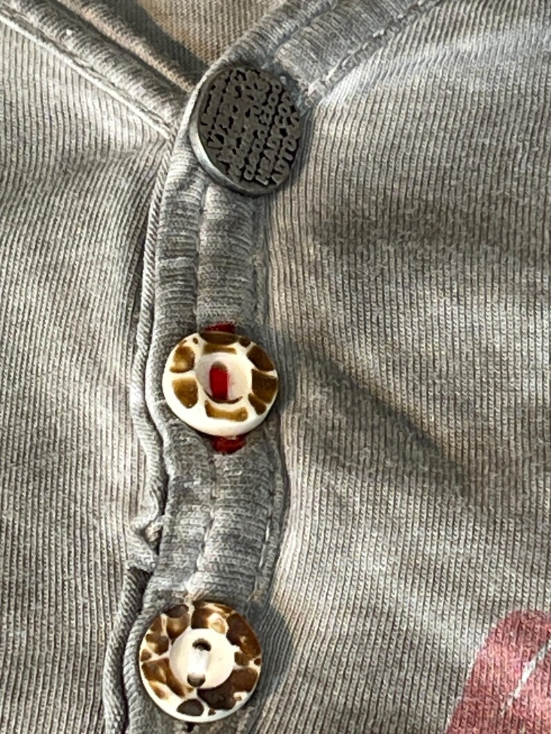 May include: Close-up of a gray ribbed knit shirt with three buttons. The top button is dark gray with embossed text. The other two buttons are white with brown and tan patterns. The shirt has a V-neck design.