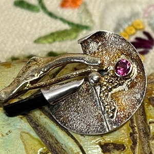 May include: A unique silver-toned brooch with a textured, circular design. It features a small, round, pink gemstone and gold-colored accents. A black, cylindrical element extends from the brooch, adding a modern touch to the artistic jewelry piece.
