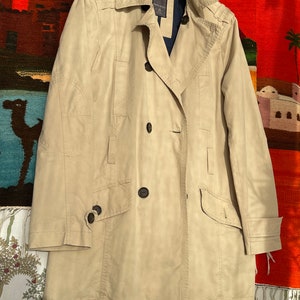 Könnte beinhalten: Ein beiger Trenchcoat mit doppelreihigem Verschluss und einer Knopfverschluss-Tasche mit Klappe auf der linken Seite. Der Mantel hat einen Kragen mit einem Knopfverschluss und einen einzigen Knopf auf der rechten Seite der Brust.