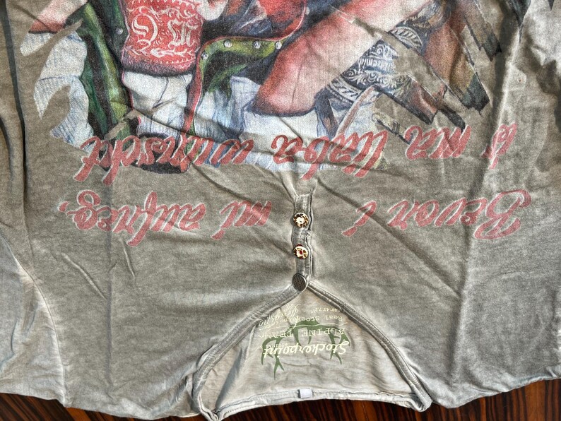 May include: A light gray Henley shirt with a graphic print and text in red. The shirt has three gold-colored buttons and a small logo at the bottom. The shirt is made of a soft, comfortable material.