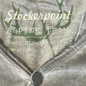 May include: Close-up of a gray Henley shirt with a button closure. The shirt features the text "Stockerpoint ALPINE TEAM" in white and green, along with a deer antler graphic. The shirt has a V-neck design.