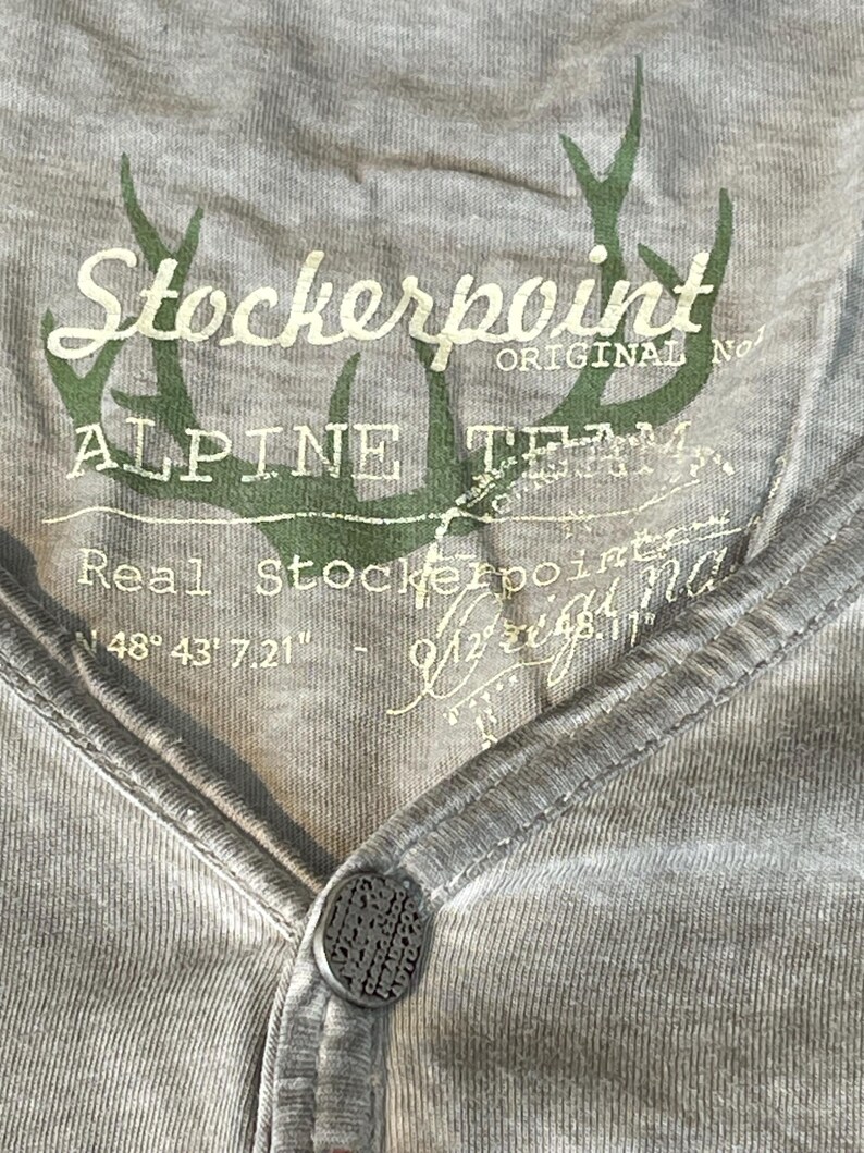 May include: Close-up of a gray Henley shirt with a button closure. The shirt features the text "Stockerpoint ALPINE TEAM" in white and green, along with a deer antler graphic. The shirt has a V-neck design.
