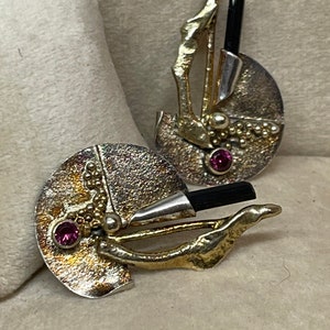 May include: Abstract silver and gold-toned earrings with a textured finish. Each earring features a black rod, a small pink gemstone, and a unique, organic design. The earrings have a post-back closure.