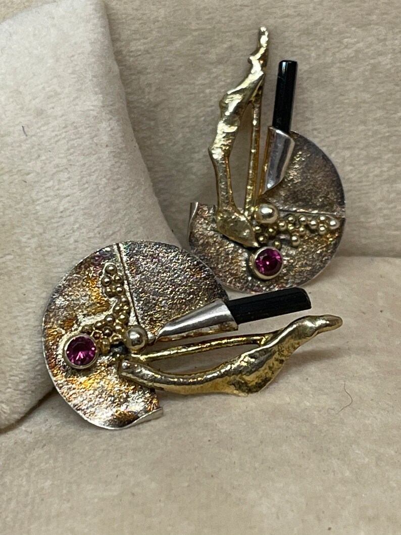 May include: Abstract silver and gold-toned earrings with a textured finish. Each earring features a black rod, a small pink gemstone, and a unique, organic design. The earrings have a post-back closure.