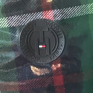 Puede incluir: Una tela de cuadros negros y verdes con un parche de cuero negro que dice "Tommy Hilfiger" en letras blancas.