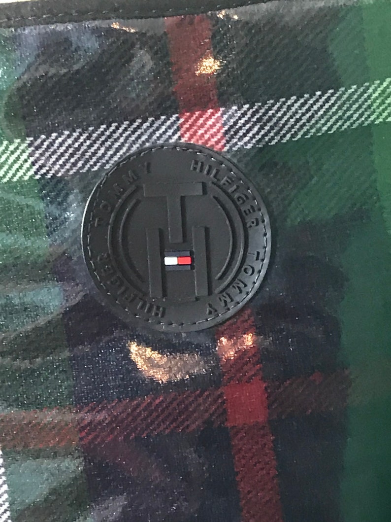 Puede incluir: Una tela de cuadros negros y verdes con un parche de cuero negro que dice "Tommy Hilfiger" en letras blancas.