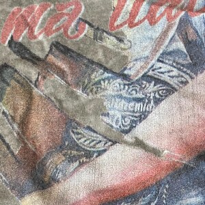 May include: Close-up of a faded olive green t-shirt with a graphic design. The design features the red text "ma love" over a faded image of a person wearing a patterned shirt and jeans.