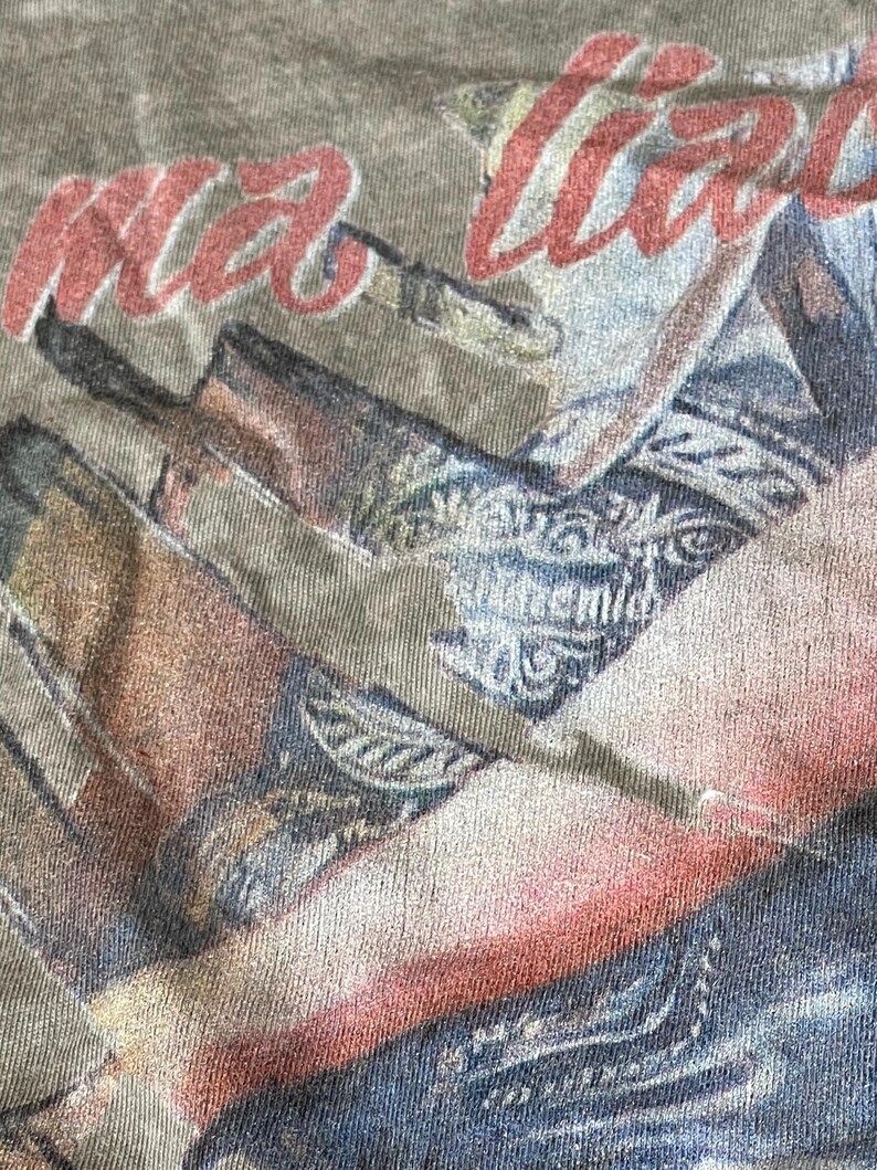 May include: Close-up of a faded olive green t-shirt with a graphic design. The design features the red text "ma love" over a faded image of a person wearing a patterned shirt and jeans.
