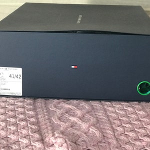 Puede incluir: Una caja de zapatos negra con el logotipo de Tommy Hilfiger y la talla 41/42 impresa en el lateral. La caja tiene un c&iacute;rculo verde en el lado derecho.