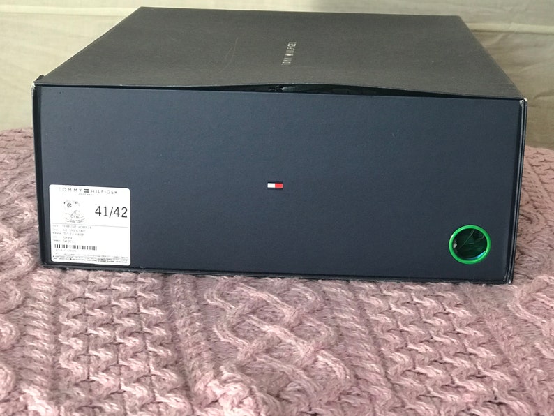 Puede incluir: Una caja de zapatos negra con el logotipo de Tommy Hilfiger y la talla 41/42 impresa en el lateral. La caja tiene un c&iacute;rculo verde en el lado derecho.
