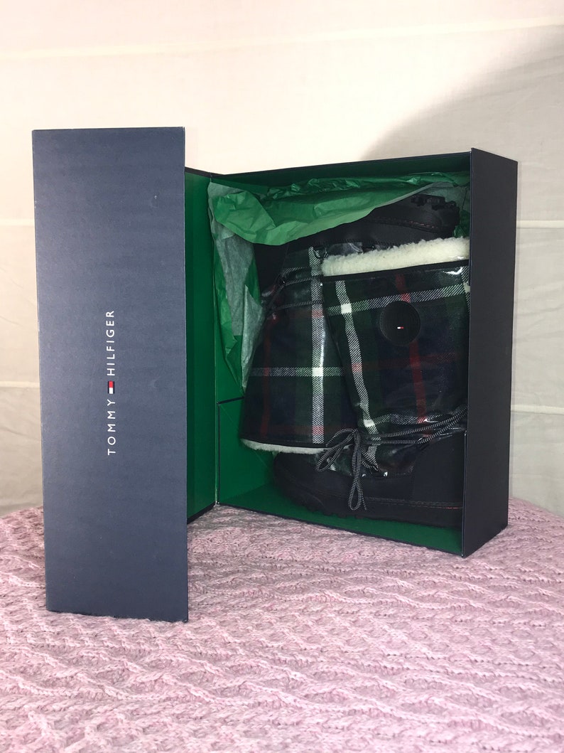 Puede incluir: Un par de botas de invierno negras con un patr&oacute;n de cuadros y un forro de piel blanca, dentro de una caja azul con el logotipo de Tommy Hilfiger.