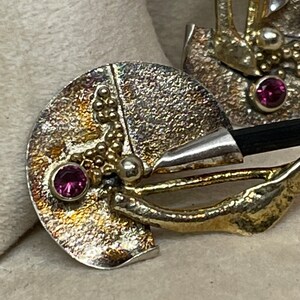 May include: Close-up of a handcrafted silver and gold-toned brooch. The brooch features a circular design with a textured surface, small gold beads, and a vibrant pink gemstone. A black element is also visible.