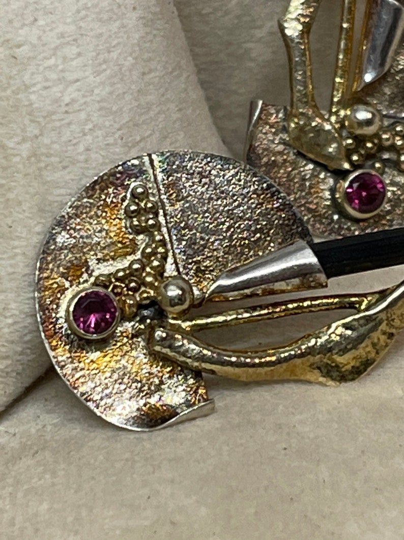 May include: Close-up of a handcrafted silver and gold-toned brooch. The brooch features a circular design with a textured surface, small gold beads, and a vibrant pink gemstone. A black element is also visible.