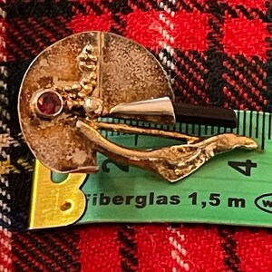 May include: A vintage silver-toned brooch with a circular design, featuring a small red gemstone and a gold-colored, leaf-like accent. The brooch is set against a red and black plaid background.