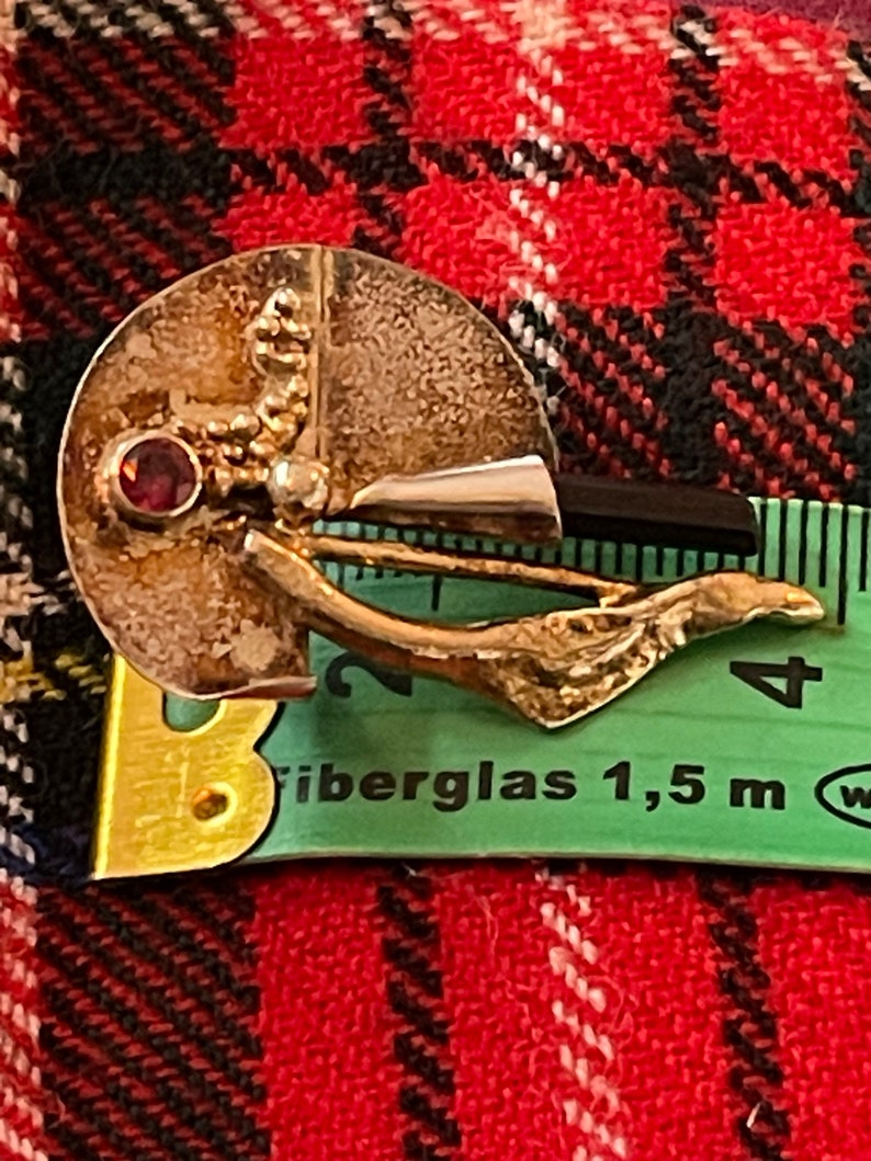 May include: A vintage silver-toned brooch with a circular design, featuring a small red gemstone and a gold-colored, leaf-like accent. The brooch is set against a red and black plaid background.
