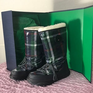 Puede incluir: Un par de botas de invierno negras y verdes a cuadros con forro de piel blanca y un detalle de bot&oacute;n negro. Las botas est&aacute;n en una caja con forro verde.
