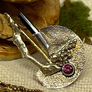 May include: A silver-toned brooch with a textured, semi-circular design. It features a black rod, a small silver ball, and a cluster of tiny silver beads. A round, pink gemstone is set near the edge. The brooch has a unique, artistic style.