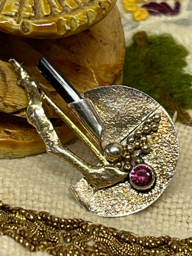 May include: A silver-toned brooch with a textured, semi-circular design. It features a black rod, a small silver ball, and a cluster of tiny silver beads. A round, pink gemstone is set near the edge. The brooch has a unique, artistic style.