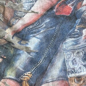 May include: Close-up of a faded graphic tee featuring a detailed illustration. The design includes elements of denim, red, and tan hues, with visible stitching and a textured appearance. The overall aesthetic suggests a vintage or worn style.