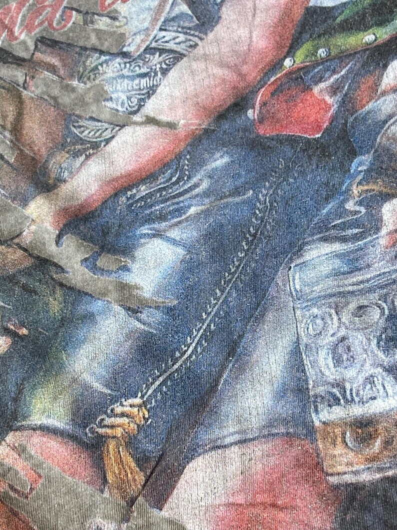 May include: Close-up of a faded graphic tee featuring a detailed illustration. The design includes elements of denim, red, and tan hues, with visible stitching and a textured appearance. The overall aesthetic suggests a vintage or worn style.
