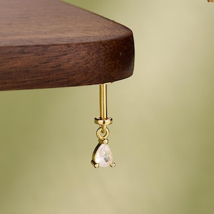 May include: A gold earring with a clear, pear-shaped gemstone hangs from a dark brown wooden surface. The word "JEW SEEN" is visible at the bottom of the image. The background is a soft green.
