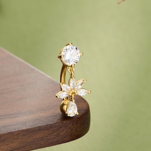 May include: A gold-colored belly button ring with a curved barbell design. The ring features a large, clear, round-cut gemstone at the top, a flower-shaped charm with multiple clear gemstones, and a teardrop-shaped gemstone at the bottom. The word "JEWSEEN" is at the bottom.
