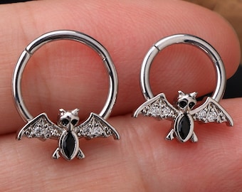 Puede incluir: Dos pendientes de aro plateados con un diseño de murciélago. Cada murciélago tiene ojos negros y alas con pequeñas piedras transparentes.
