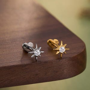 May include: Two starburst stud earrings, one silver and one gold, each with a central clear gemstone. The earrings are displayed on a dark wooden surface. The word "JEWSEEN" is printed at the bottom.