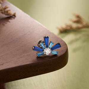 Puede incluir: Un pendiente de botón plateado con un diseño de explosión. El pendiente presenta una piedra central transparente, rodeada de piedras azules y celeste talladas en baguette. La palabra "JEWSEEN" está en la parte inferior de la imagen.