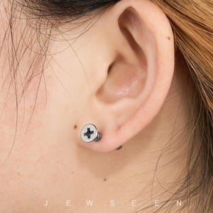 May include: A close-up of an ear with a silver screw-shaped earring. The earring has a white head with a black cross design. The word "JEWSEN" is printed at the bottom of the image.