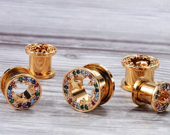 May include: Four gold-colored ear gauges with a star-shaped design and colorful gemstones. The gauges are arranged on a light wood surface.