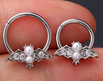 Puede incluir: Dos anillos de septum plateados con un diseño de murciélago y detalles de perlas. El murciélago tiene diamantes de imitación transparentes alrededor de las alas.