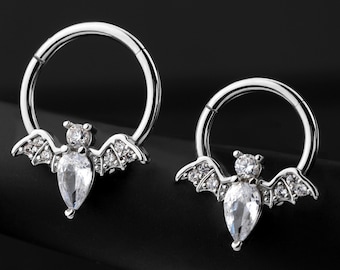 Puede incluir: Dos anillos de septum de plata con dijes de murciélago de cristal transparente. El anillo más grande está a la izquierda y el más pequeño a la derecha. Los anillos están sobre un fondo negro.