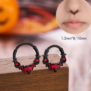 以下が含まれることがあります： 赤いハート型の宝石とアクセントが付いた黒いノーズリング。リングは木製の表面に表示され、下部に「JEWSEEN」という文字があります。鼻ピアスをクローズアップしたものが右上隅に表示されています。