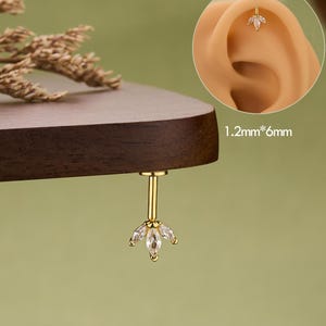 Peut inclure: Un piercing d'oreille doré avec un motif floral, orné de pierres précieuses transparentes. Le piercing est présenté en détail, avec une vue rapprochée de l'oreille. Le texte "JEWSEEN" est au bas de l'image.