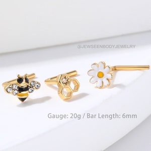 May include: Three gold nose rings with different designs: a bee with black and yellow stripes and clear rhinestones, a honeycomb with a clear rhinestone, and a white daisy with a yellow center. Gauge: 20g / Bar Length: 6mm.