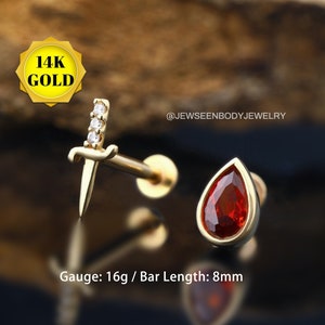 Op de afbeelding: Twee goudkleurige oorbellen. Eén is een dolkvorm met drie heldere stenen. De andere is een druppelvorm met een rode steen. De tekst op de afbeelding luidt "14K GOLD" en "@JEWSEENBODYJEWELRY". De gauge is 16g en de staaf lengte is 8mm.
