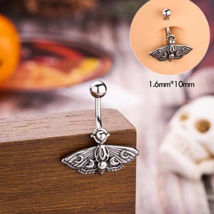 May include: A silver belly button ring with a moth pendant. The moth has detailed wing patterns and is suspended from a curved barbell with a small ball at the top. The text "1.6mm*10mm" is visible.