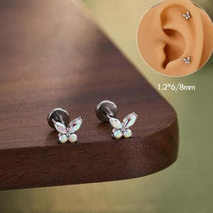 May include: Two butterfly-shaped ear studs with iridescent crystals. The studs are silver-colored and have a flat back. The image also shows a close-up of an ear with the studs in place. The text "JEWSEEN" is at the bottom.