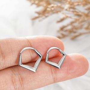 Puede incluir: Un par de pendientes de aro geométricos pequeños de color plateado. Los pendientes tienen un diseño angular único, con forma de pentágono. Los pendientes se muestran sobre un fondo claro, con la palabra "JEWSEEN" visible en la parte inferior.