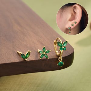 May include: Gold-toned earrings with green gemstone accents. The set includes a butterfly stud, a flower stud, and a hoop earring with a dangling gem. The earrings are displayed on a wooden surface. The word "JEWSEEN" is printed at the bottom.