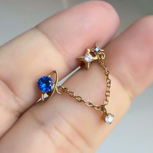 Könnte beinhalten: Ein goldfarbener Kettenohrring mit einem blauen runden Stein und einem umgebenden Ring, der über eine feine Gliederkette mit einem sternförmigen Anhänger verbunden ist, welcher einen klaren und einen kleinen blauen Stein sowie einen klaren Stein unten hat.