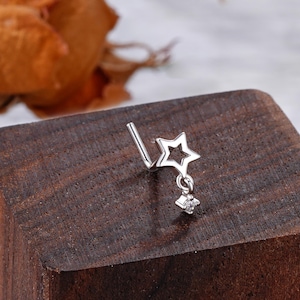 Puede incluir: Un aro de nariz de plata con un colgante de estrella y una pequeña piedra transparente. El aro está sobre una superficie de madera.