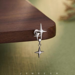 Puede incluir: Un pendiente plateado con diseño de estrella. El pendiente presenta tres estrellas, cada una con una gema transparente en el centro. Las estrellas están conectadas por una delicada cadena. La palabra "JEWSEEN" está impresa en la parte inferior de la imagen.